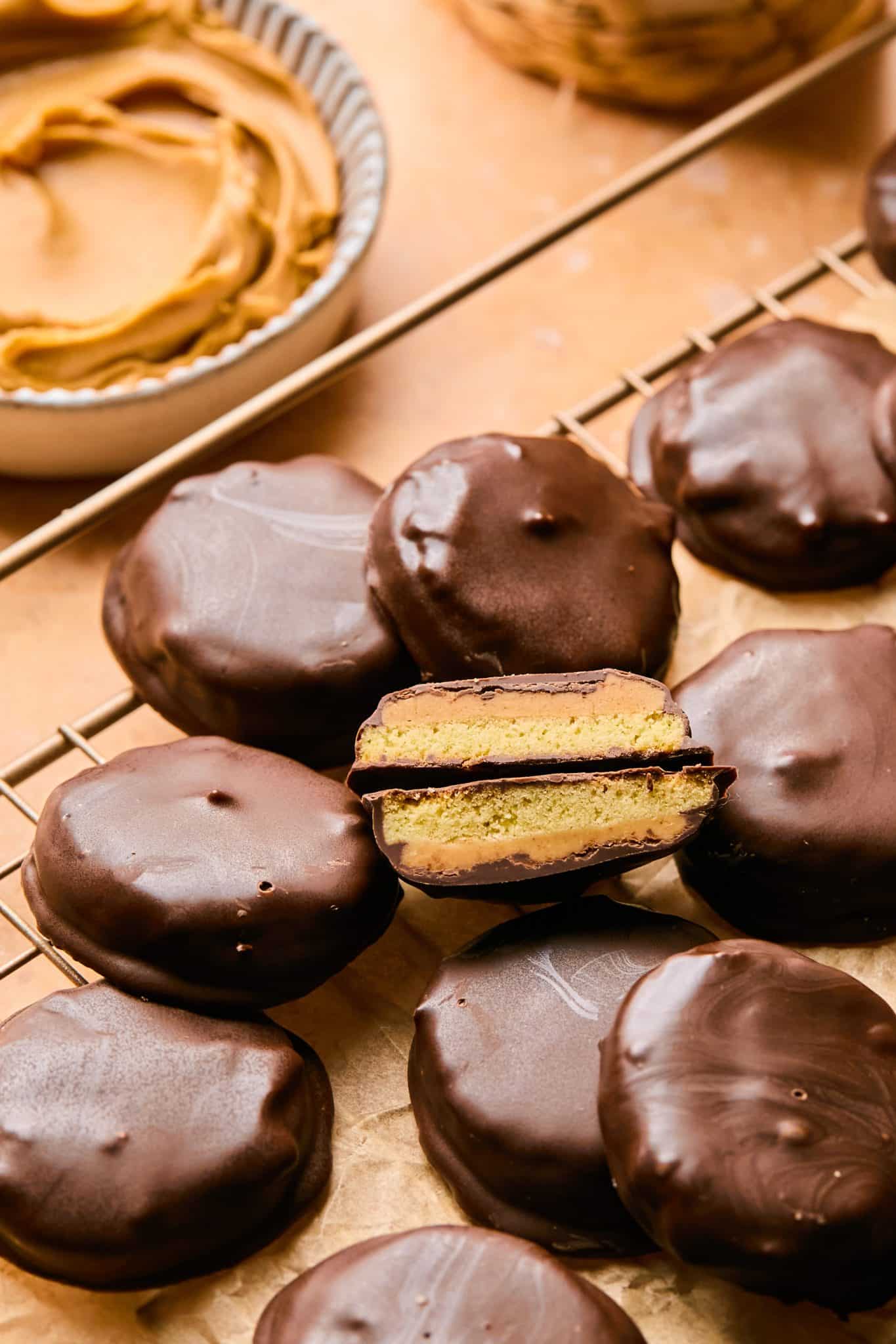 A wire rack filled with chocolate-coated Tagalong copycat cookies, with one cookie sliced in half to reveal a golden peanut butter layer sandwiched between the cookie base and the chocolate coating, and a small bowl of peanut butter in the background.



